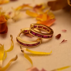 May include: Two gold hoop earrings with a row of rectangular, deep pink gemstones. The earrings are set against a soft, neutral background with scattered yellow and orange flower petals, creating a delicate and elegant aesthetic.