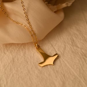 Könnte beinhalten: Eine goldfarbene Halskette mit einem polierten, glatten Anhänger in Form eines stilisierten Hammers. Der Anhänger hängt an einer zarten, dünnen goldfarbenen Kette mit kleinen, gleichmäßigen runden Gliedern, was ein schlichtes, reflektierendes Aussehen ergibt.