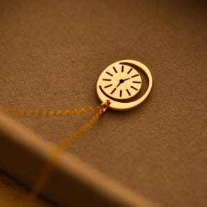 May include: A gold-colored pendant necklace featuring a clock design. The clock face is inside an oval shape with a crescent moon cutout. The necklace chain is also gold. The design is simple and elegant.