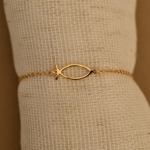 Puede incluir: Una pulsera dorada con un colgante con forma de pez. La cadena y el colgante de pez son de metal amarillo brillante. El símbolo del pez es un contorno abierto. La pulsera se muestra sobre un fondo neutro.