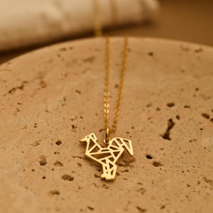 Può includere: Una collana color oro con un ciondolo a forma di gallo geometrico. Il ciondolo è composto da forme triangolari e quadrilatere interconnesse. La catena è delicata e pende dal gallo. La collana è esposta su una superficie strutturata marrone chiaro.