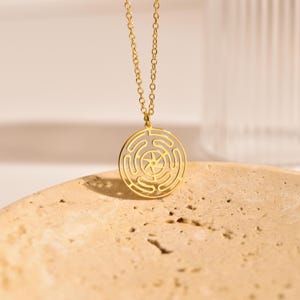 Peut inclure: Un collier pendentif doré avec un motif circulaire en forme de labyrinthe et une étoile au centre. La chaîne est fine. Le pendentif est présenté sur une surface texturée de couleur brun clair.