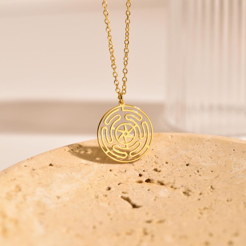 May include: A gold-colored pendant necklace featuring a circular design with a maze-like pattern and a star in the center. The necklace hangs from a delicate chain. The pendant is displayed on a textured, light-brown surface.