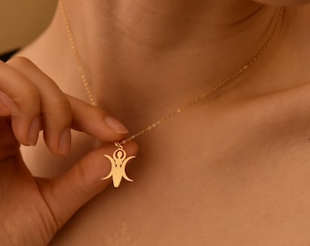 May include: A gold-colored necklace with a pendant featuring a stylized figure with outstretched arms and a crescent moon above. The delicate chain is held by a hand, showcasing the pendant's intricate design and warm metallic hue.