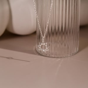 Può includere: Una collana d'argento con un ciondolo a forma di stella geometrica. La stella è realizzata in metallo sottile e ha un centro vuoto.