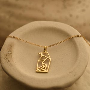 May include: A gold necklace with a geometric penguin pendant. The pendant is a stylized outline of a penguin. The necklace is displayed on a neutral-toned surface. The chain is delicate.