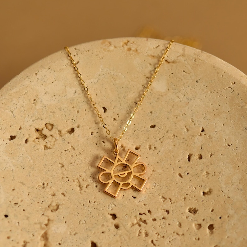 May include: A gold-colored necklace with a pendant shaped like a stylized sun. The pendant has a geometric design with a smiling face in the center. The necklace is displayed on a beige, textured surface.