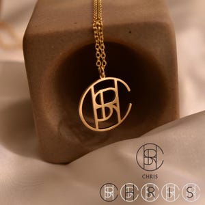 May include: A gold-coloured pendant necklace with the letters "CB" inside an oval. The chain is fine and delicate. The word "CHRIS" is printed below the pendant in a minimalist font. The necklace is displayed against a neutral background.