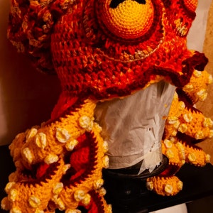 Pode incluir: Um chap&eacute;u de polvo de croch&ecirc; com fio vermelho, laranja e amarelo. O chap&eacute;u tem um olho grande com uma pupila preta e uma boca preta.