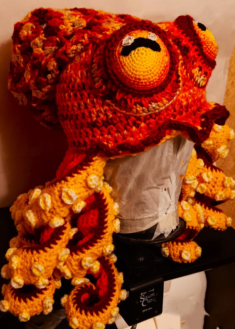 Pode incluir: Um chap&eacute;u de polvo de croch&ecirc; com fio vermelho, laranja e amarelo. O chap&eacute;u tem um olho grande com uma pupila preta e uma boca preta.