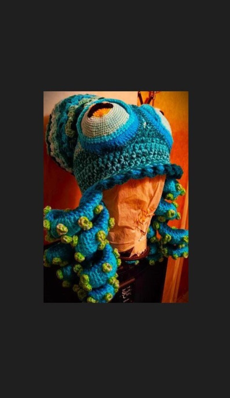Pode incluir: Um chap&eacute;u de polvo de croch&ecirc; azul e verde com um olho grande no topo. O chap&eacute;u &eacute; usado em uma cabe&ccedil;a de manequim.