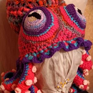 Pode incluir: Um chap&eacute;u de polvo de croch&ecirc; com fio rosa, roxo e azul. O chap&eacute;u tem uma cabe&ccedil;a grande e redonda com dois olhos grandes e oito tent&aacute;culos.