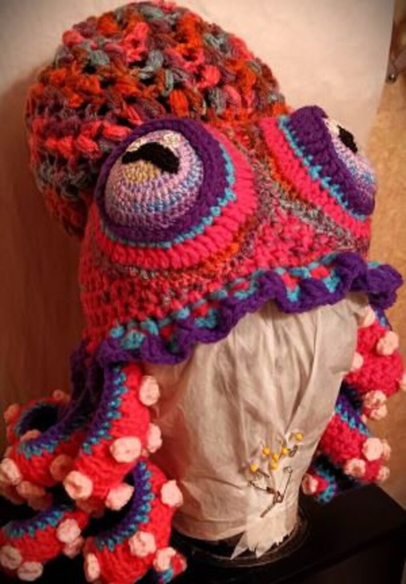 Pode incluir: Um chap&eacute;u de polvo de croch&ecirc; com fio rosa, roxo e azul. O chap&eacute;u tem uma cabe&ccedil;a grande e redonda com dois olhos grandes e oito tent&aacute;culos.