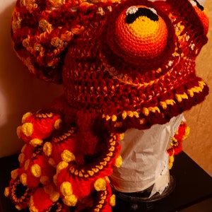 Pode incluir: Um chap&eacute;u de polvo de croch&ecirc; feito com fio vermelho, laranja e amarelo. O chap&eacute;u tem uma cabe&ccedil;a grande com dois olhos e tent&aacute;culos que est&atilde;o enrolados.