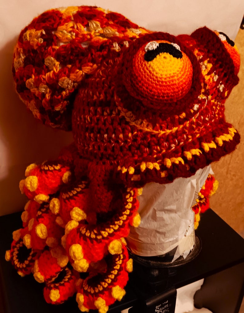 Pode incluir: Um chap&eacute;u de polvo de croch&ecirc; feito com fio vermelho, laranja e amarelo. O chap&eacute;u tem uma cabe&ccedil;a grande com dois olhos e tent&aacute;culos que est&atilde;o enrolados.