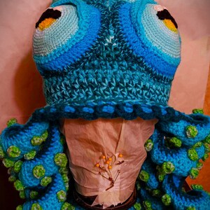 Pode incluir: Um chap&eacute;u de polvo de croch&ecirc; feito com fio azul e verde. O chap&eacute;u tem olhos grandes e tent&aacute;culos.