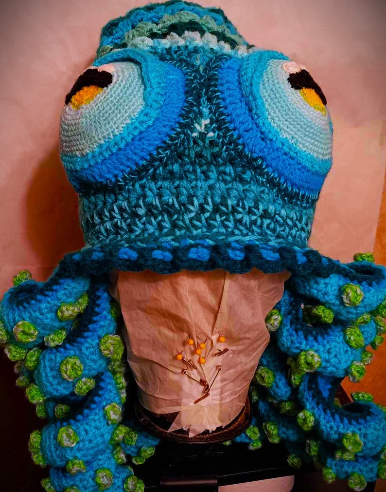 Pode incluir: Um chap&eacute;u de polvo de croch&ecirc; feito com fio azul e verde. O chap&eacute;u tem olhos grandes e tent&aacute;culos.