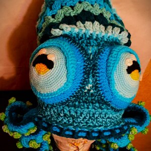 Pode incluir: Um chap&eacute;u de polvo de croch&ecirc; em tons de azul e verde. O chap&eacute;u tem olhos grandes e redondos e um corpo texturizado de croch&ecirc;. O polvo tem oito tent&aacute;culos.