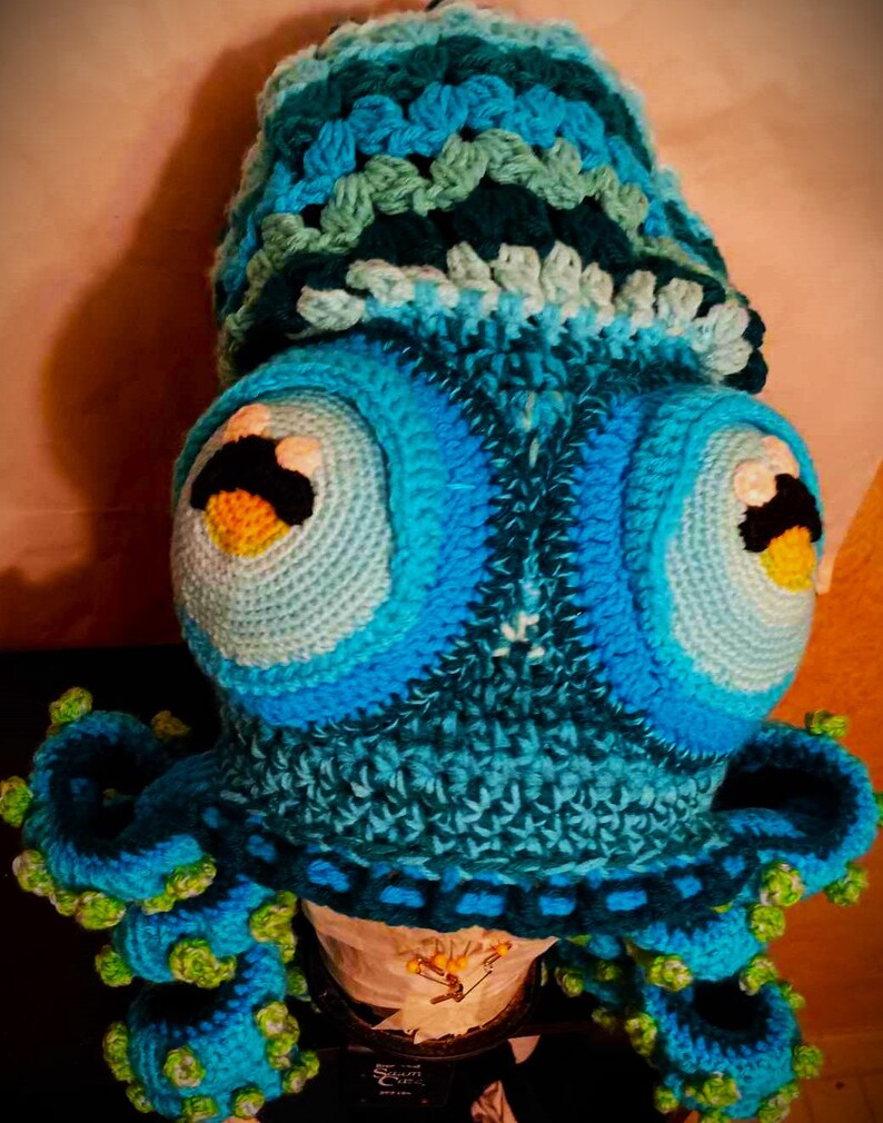 Pode incluir: Um chap&eacute;u de polvo de croch&ecirc; em tons de azul e verde. O chap&eacute;u tem olhos grandes e redondos e um corpo texturizado de croch&ecirc;. O polvo tem oito tent&aacute;culos.