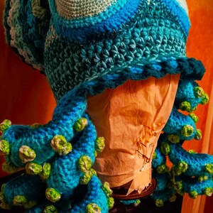 Pode incluir: Um chap&eacute;u de polvo de croch&ecirc; feito com fio azul e verde. O chap&eacute;u tem um olho grande com uma pupilla amarela e preta. Os tent&aacute;culos s&atilde;o enrolados e t&ecirc;m detalhes verdes.