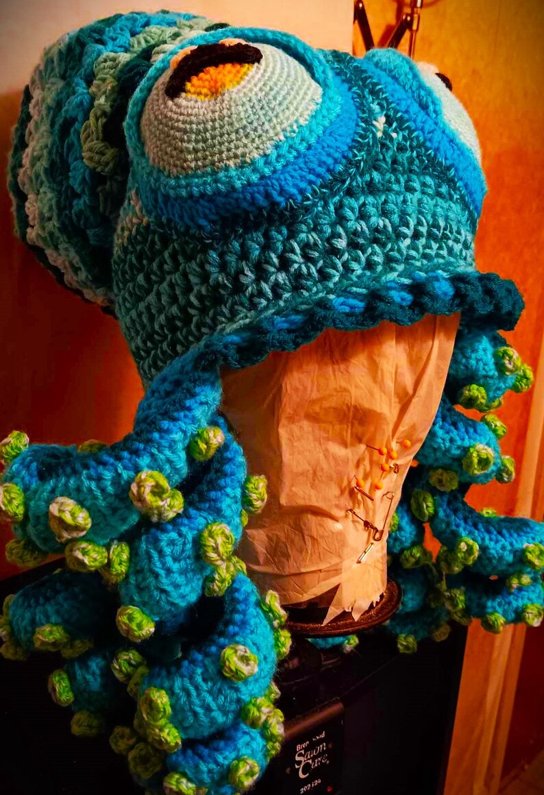 Pode incluir: Um chap&eacute;u de polvo de croch&ecirc; feito com fio azul e verde. O chap&eacute;u tem um olho grande com uma pupilla amarela e preta. Os tent&aacute;culos s&atilde;o enrolados e t&ecirc;m detalhes verdes.