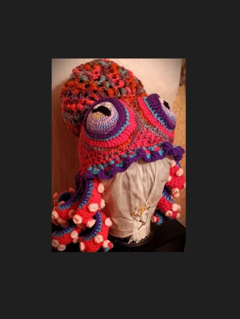 Pode incluir: Um chap&eacute;u de polvo de croch&ecirc; feito com fio rosa, roxo e azul. O chap&eacute;u tem olhos grandes e tent&aacute;culos.