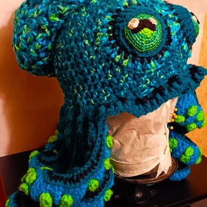 Pode incluir: Um chap&eacute;u de polvo de croch&ecirc; feito com fio azul, verde e preto. O chap&eacute;u tem olhos grandes e redondos e tent&aacute;culos com detalhes verdes e azuis.