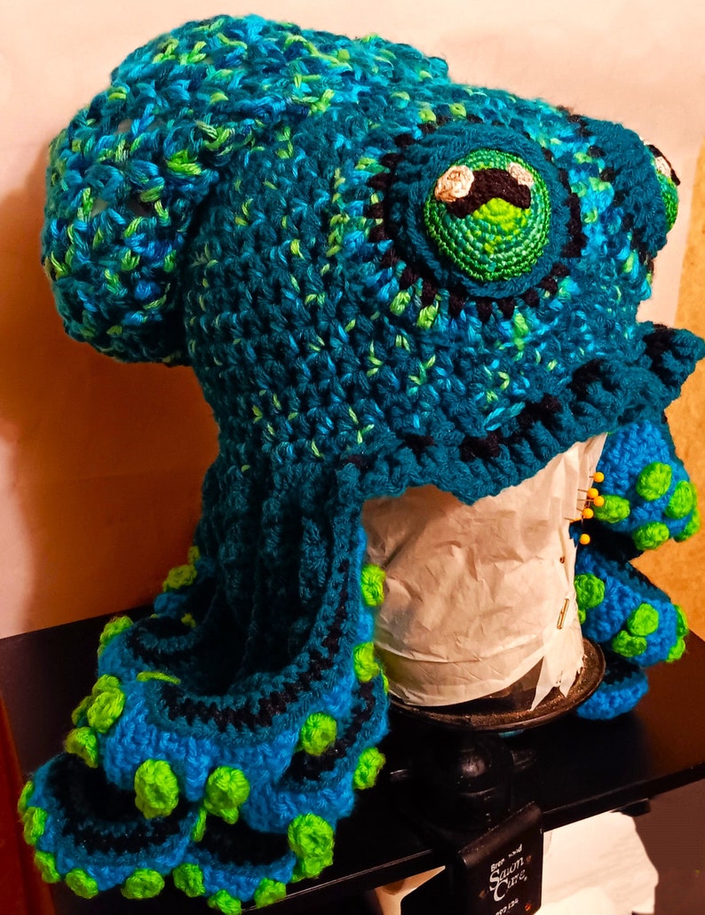Pode incluir: Um chap&eacute;u de polvo de croch&ecirc; feito com fio azul, verde e preto. O chap&eacute;u tem olhos grandes e redondos e tent&aacute;culos com detalhes verdes e azuis.