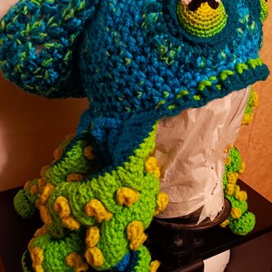 Pode incluir: Um chap&eacute;u de polvo de croch&ecirc; com um corpo azul e verde e tent&aacute;culos amarelos. O chap&eacute;u est&aacute; em uma cabe&ccedil;a de manequim.