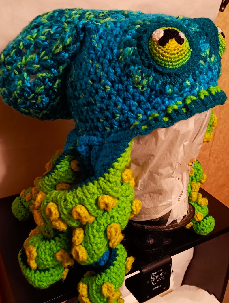 Pode incluir: Um chap&eacute;u de polvo de croch&ecirc; com um corpo azul e verde e tent&aacute;culos amarelos. O chap&eacute;u est&aacute; em uma cabe&ccedil;a de manequim.