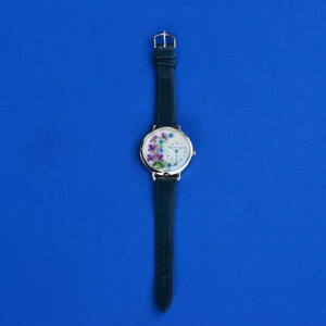 May include: A silver wristwatch with a white face and a green leather band. The face of the watch has a floral design with purple, pink, and blue flowers.