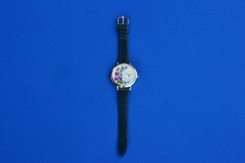 May include: A silver wristwatch with a white face and a green leather band. The face of the watch has a floral design with purple, pink, and blue flowers.