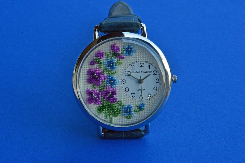 May include: A silver wristwatch with a white face and black hands. The watch face has a floral design with purple and blue flowers. The watch face also has the text "Elizabeth H Anderson LONDON" printed on it.