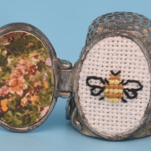 Puede incluir: Un dedal de plata con una tapa con bisagras. La tapa tiene una imagen de un jardín de flores. El dedal tiene un diseño de punto de cruz de una abeja en la parte delantera.