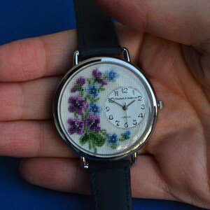 May include: A silver wristwatch with a white face and a floral design. The watch face has black Roman numeral hour markers and black hands. The watch band is black leather.