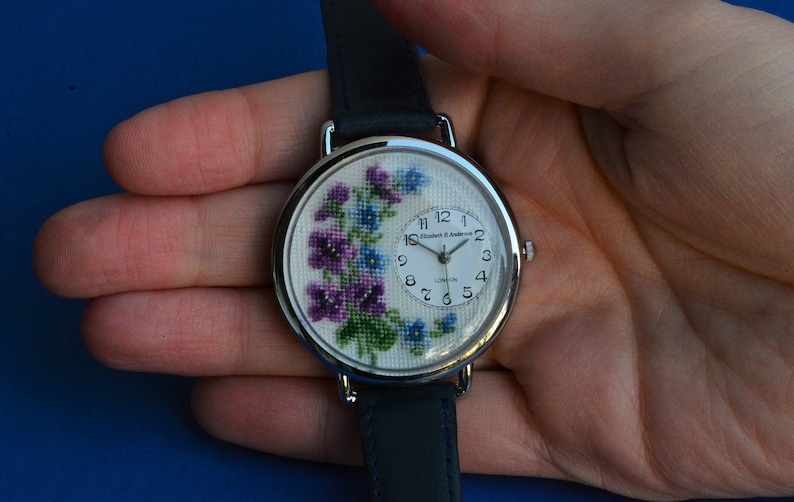 May include: A silver wristwatch with a white face and a floral design. The watch face has black Roman numeral hour markers and black hands. The watch band is black leather.