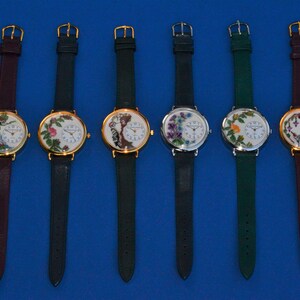 May include: Six wristwatches with different floral embroidery designs on the face. The watches have gold, silver, and white face plates with black hands. The bands are either dark green or burgundy.