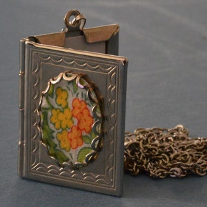 Può includere: Una collana con un ciondolo d'argento, con un designo di tessuto floreale all'interno. Il ciondolo è aperto e la catena è visibile.
