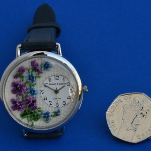 May include: A silver wristwatch with a white face and a floral design. The watch has a black leather band and a small coin next to it.