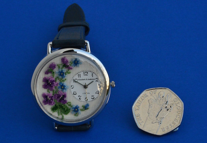 May include: A silver wristwatch with a white face and a floral design. The watch has a black leather band and a small coin next to it.