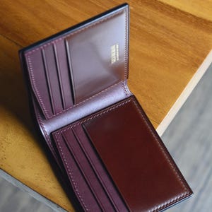 Puede incluir: Cartera de cuero marrón abierta, con interior burdeos. La cartera tiene múltiples ranuras para tarjetas y un compartimento para billetes. Los bordes están cuidadosamente cosidos y la cartera está sobre una superficie de madera.