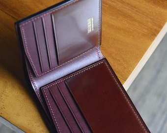 May include: Open, brown leather wallet with a burgundy interior. The wallet has multiple card slots and a bill compartment. The edges are neatly stitched, and the wallet is set against a wooden surface.
