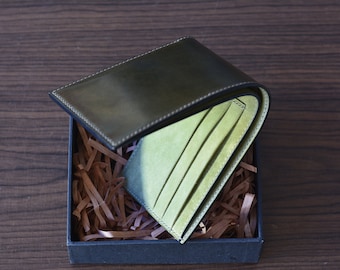 May include: A green leather wallet with a black leather flap, folded open to reveal a green interior with multiple card slots. The wallet is sitting in a box filled with brown packing paper.