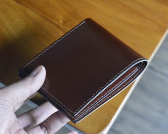 May include: A brown leather wallet with white stitching around the edges. The wallet is rectangular and appears to be folded. The image is taken from a slightly elevated angle, showing the top of the wallet.