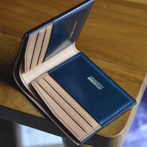 Könnte beinhalten: Ein aufklappbares Portemonnaie mit dunkelblauer Außenseite und beigefarbener Innenseite mit mehreren Kartenfächern. Das Innere ist mit dem Text Gitsi Roshani Leatherworks in goldener Schrift geprägt und weist sichtbare Nähte auf.