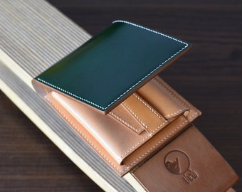 May include: A handcrafted leather wallet with a dark green cover and tan interior. The wallet features white stitching and multiple card slots. The design includes a small embossed logo on the exterior. The wallet is displayed on a wooden surface.