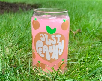 May include: A pink glass with a peach design and the text "Just Peachy" on it. The glass is filled with a pink beverage and is sitting on green grass.