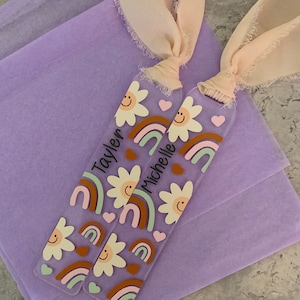May include: Two personalized acrylic bookmarks with rainbow and daisy designs. The bookmarks are tied with pink ribbon and the names "Taylen" and "Michelle" are written on them.