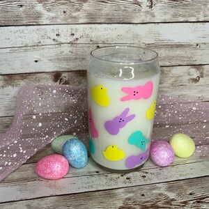 Op de afbeelding: Een heldere glazen kaars met een witte was binnenkant, versierd met kleurrijke paasvormen. De kaars is omgeven door pastelkleurige paaseieren en een roze lint op een houten oppervlak. De vormen omvatten gele kuikens, roze en paarse konijntjes en turquoise vormen.
