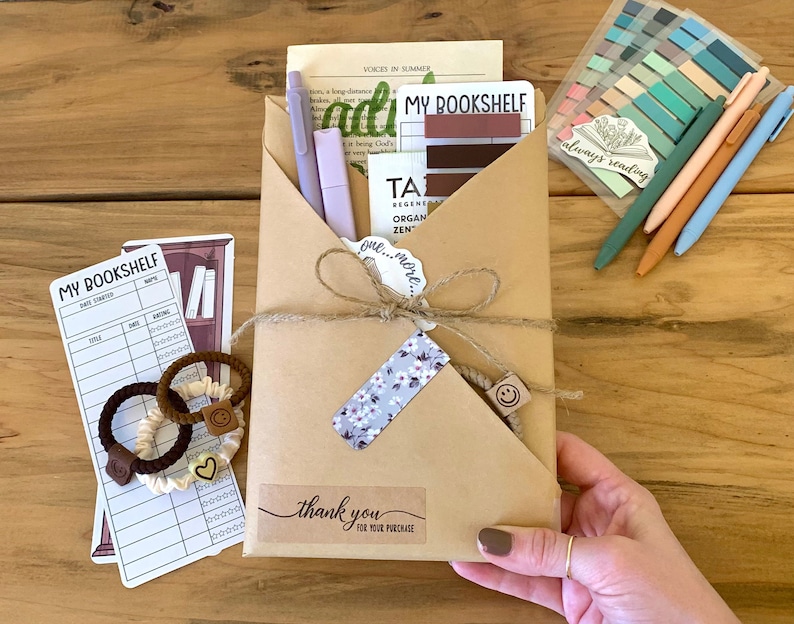 May include: A brown paper envelope with a thank you tag, tied with twine and decorated with a floral sticker. The envelope contains a variety of items, including a book tracker, pens, and hair ties. The text on the envelope reads "thank you for your purchase".